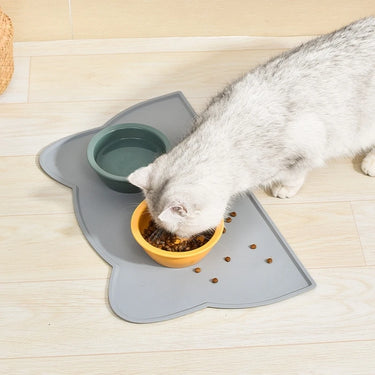 A white cat eats from a yellow Silicone Pet Feeding Bowl by KittyNook Cat Co. on a mat with spilled kibble nearby.